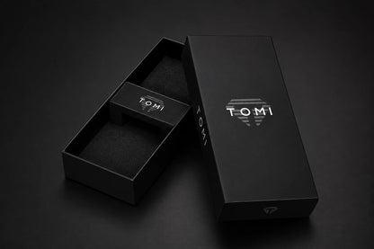 A black rectangular TOMI product box shown open with its lid leaning against it on a dark studio background. The box interior and lid are matte black with white TOMI branding and a small diamond-like logo near the bottom.